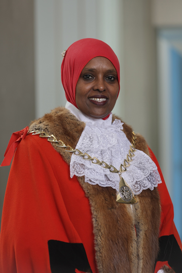 A woman wearing a cape and chain of office and a yellow headscarf